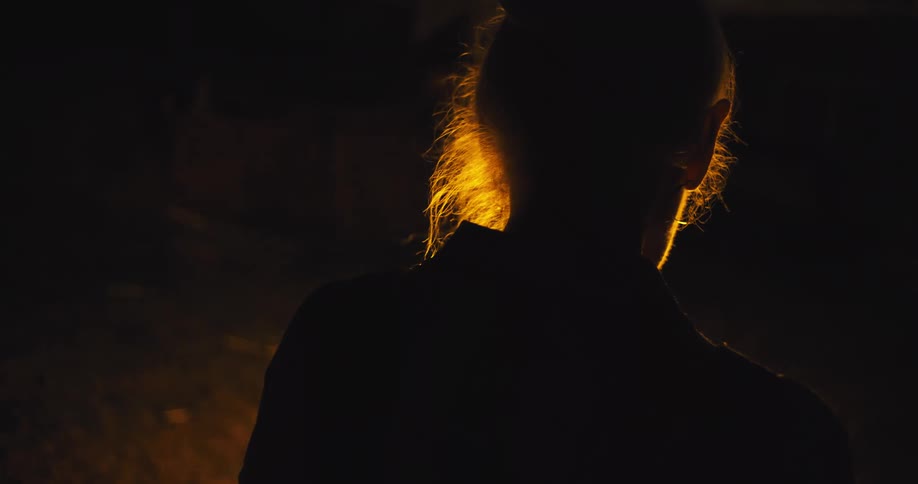 A person with long hair tied back is seen from behind their silhouette glowing in the warm light of a fire against the dark night