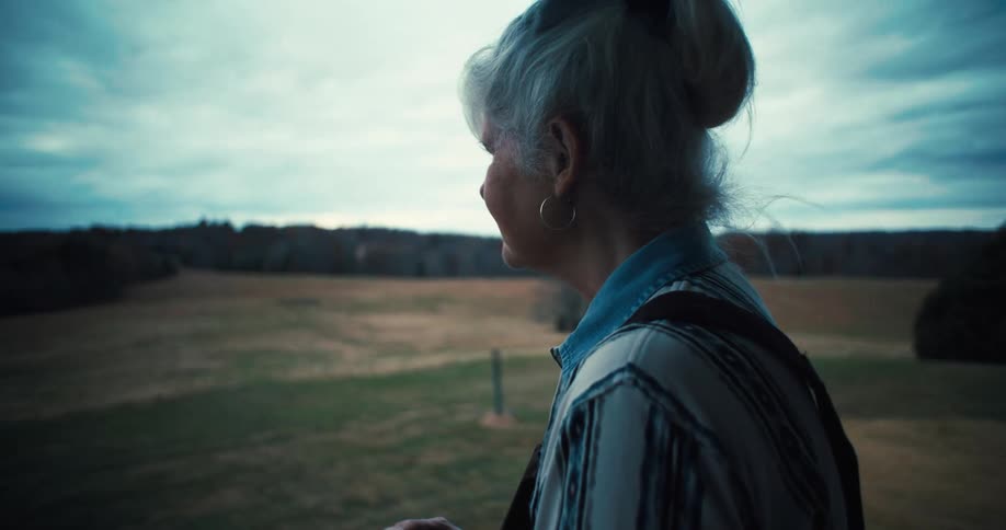 An elderly woman stands on her front porch watching over the large stretch of grassland surrounding the building at midday