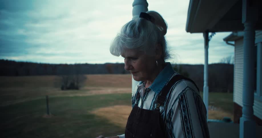 Older woman standing on the porch of her rural home holds a mug and thinks