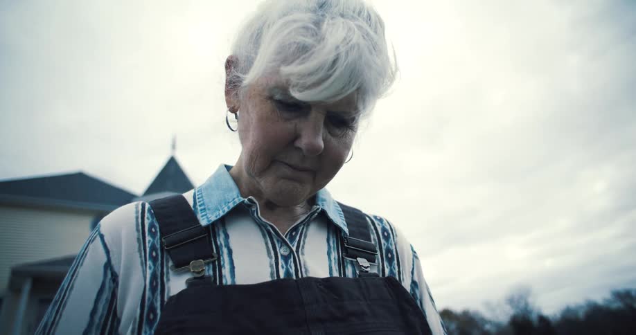 An elderly woman with gray hair stands outdoors looking sad on a cloudy day