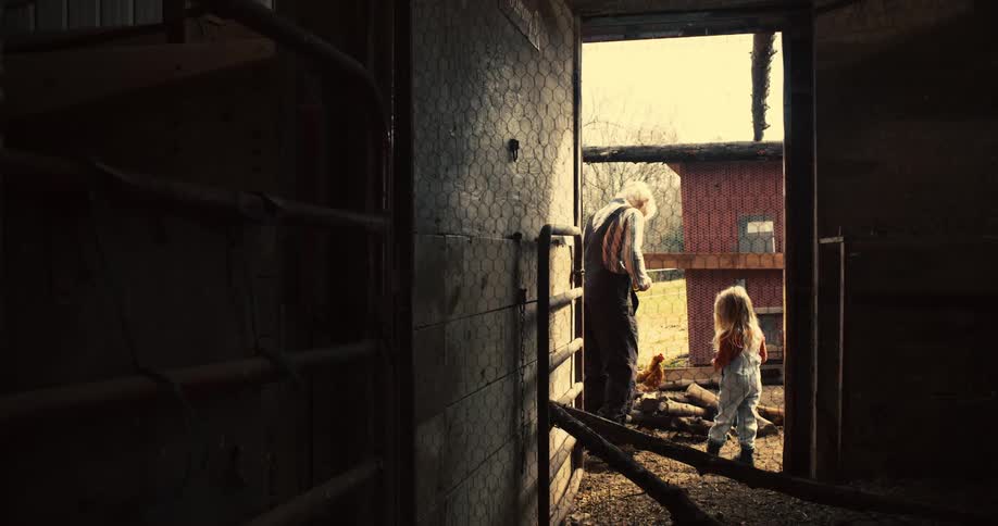 A man and a young girl stand in a sunlit farmyard near chickens framed by the doorway of a barn