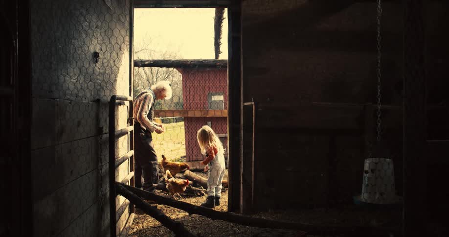 A young girl in overalls and an older woman are seen in a rural setting with the girl actively reaching into a wooden chicken coop as the woman stands beside her