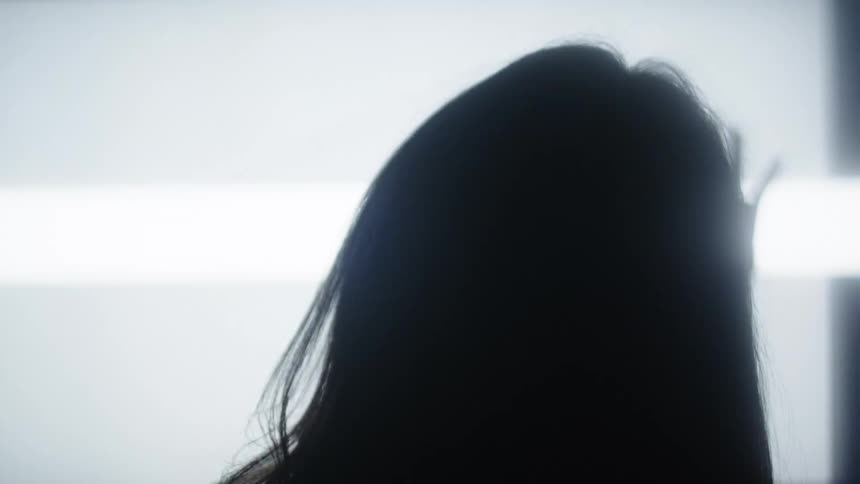 A dark silhouette of a person with long hair is visible from behind while a hands shadow presses against a white glowing surface to the right