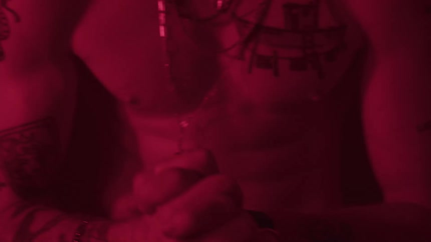 A close view of a man with many tattoos on his torso and arms is visible in dark red lighting as he holds his hands clasped together in front of him