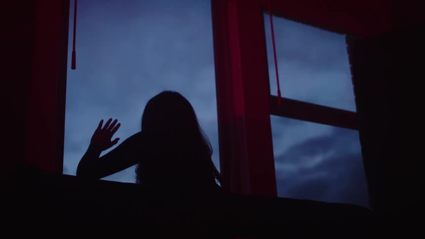A young woman standing in front of a window leaning her face against the glass as she looks outside on a cloudy evening