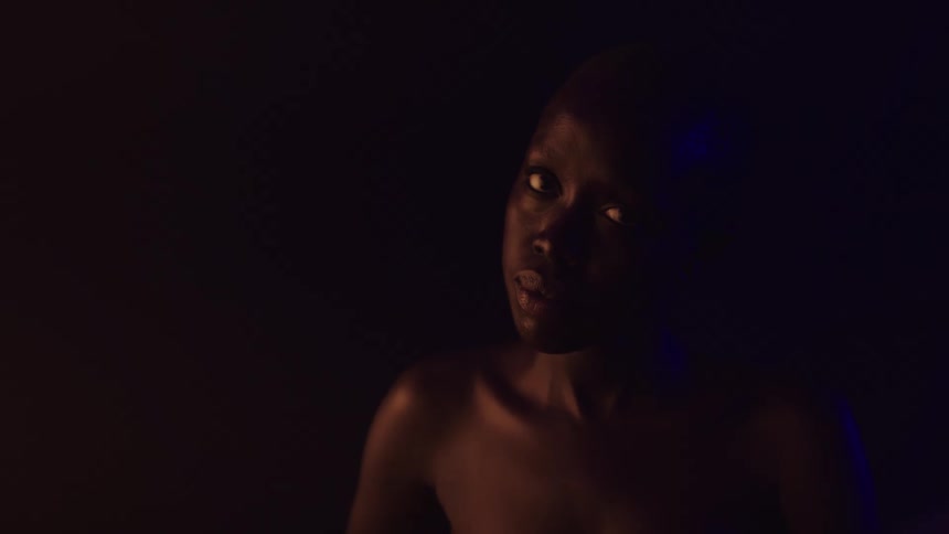 A close view of a person with dark skin shows them turning their head to the left with an orange light on the left side of their face and a blue light on the right side
