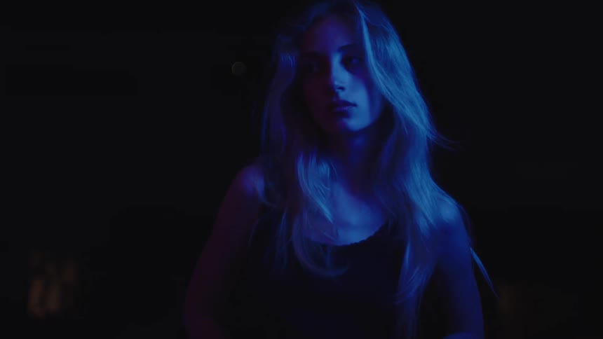 A medium shot of a woman with long light hair standing in the dark her face and hair illuminated by a combination of blue and red lights as she looks at the camera with a serious expression