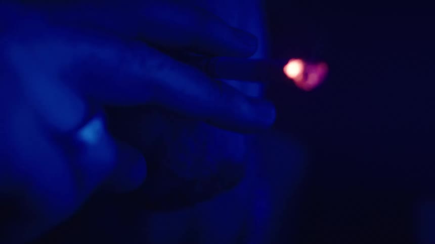 A man in a dark room illuminated by blue fluorescent light smokes a cigarette