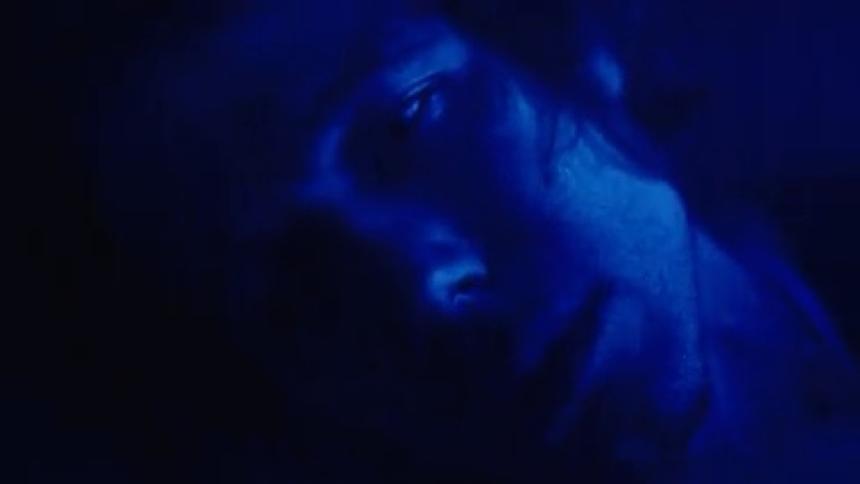 A young man sits in a dark room illuminated by dim blue light wearing a ring as he smokes a cigar