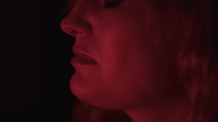 A close view of a woman with long light colored hair with her eyes closed and her head tilted up is shown as a deep red light illuminates her face