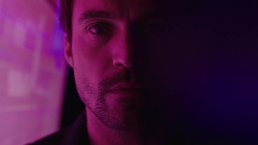 A mans face is seen in a close view with the left side illuminated by a flashing colorful screen while the right side remains in shadow as he looks directly into the camera