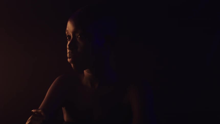 A close view shows a woman with dark skin in profile with her head turning to look over her left shoulder while a warm orange light illuminates the left side of her face