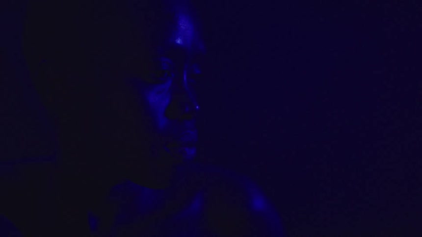 A young woman sits in a dark room illuminated by dim blue light looking sad and scared