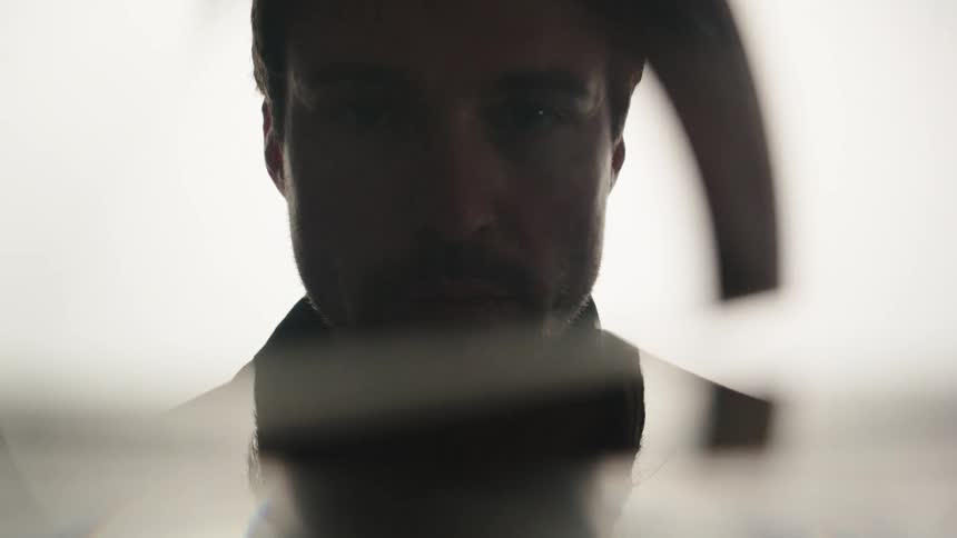 A lowangle shot shows a persons torso and head from behind a reflective or transparent surface the person is backlit by a bright light casting their front into deep shadow