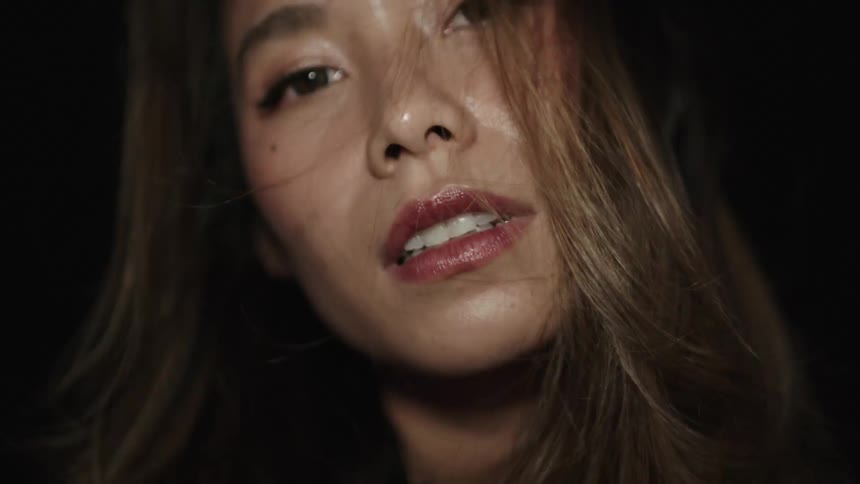 A young woman with red lipstick and long brown hair sits in a dark room her face briefly revealed as bright light flickers across it