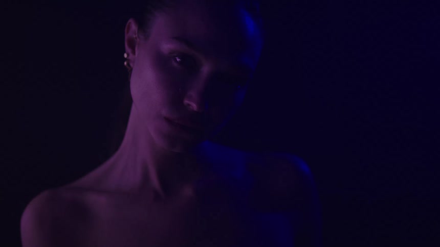 A young woman in a dark room wearing small earrings looks sad as the space is illuminated by dim blue and red light