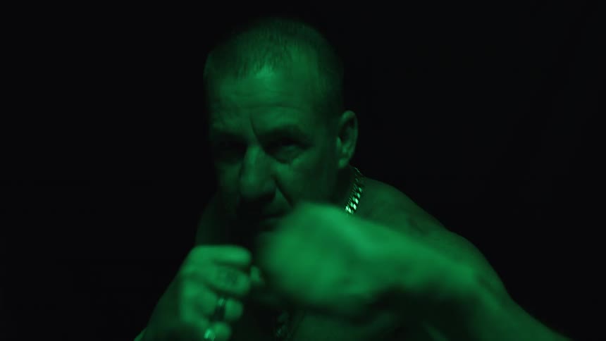 A man with short hair and a chain necklace boxes with his fists clenched while a green light illuminates his face and shoulders against a dark background
