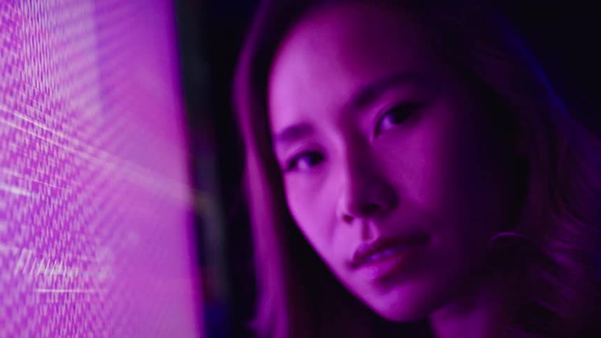 A young asian woman wearing lipstick stands in a dark room in front of a large monitor screen displaying a distorted image