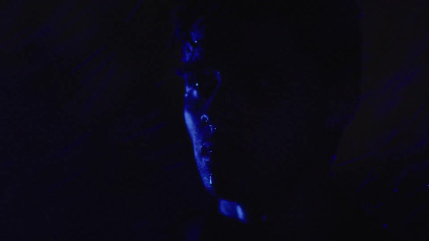 A man with an angry look in the dark with only a dim blue light falling on part of his face as rain falls on him