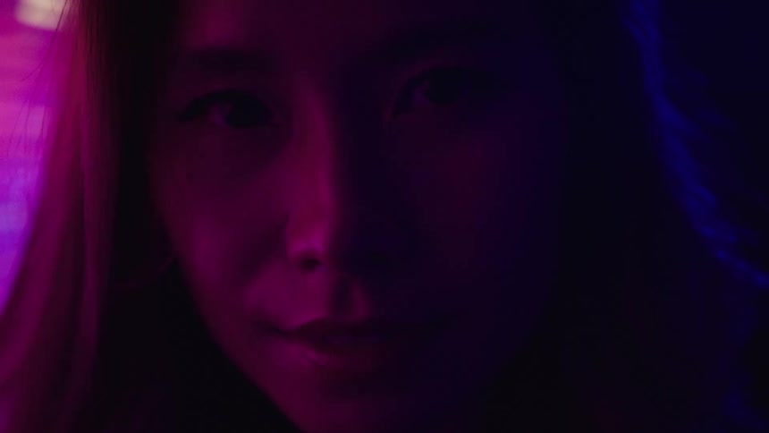 A womans face is seen in a close view with her eyes looking directly into the camera as flashing colorful light illuminates the left side of her face