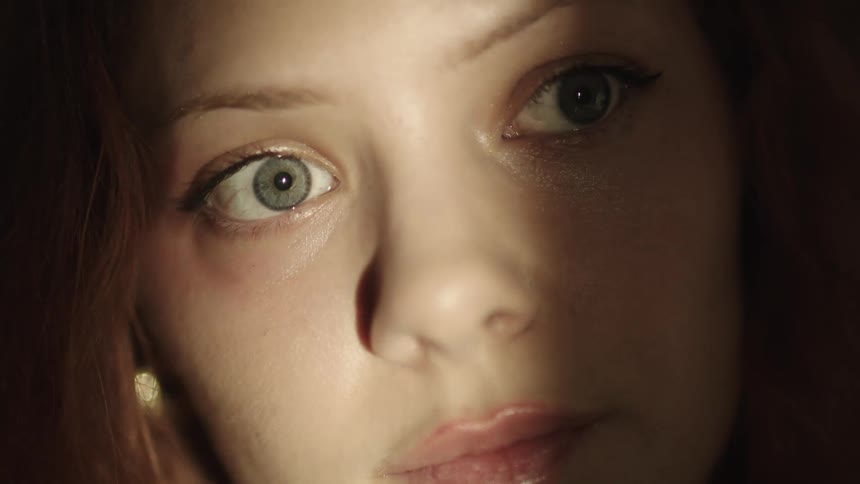 A young woman with brown hair sits inside a dark room as light moves from right to left briefly revealing her face