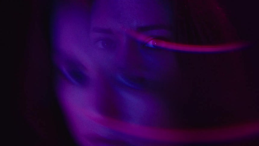 A persons face is illuminated with a strong purple light with their eyes looking directly at the camera thin blurry magenta streaks or lines pass across the frame creating a distorted effect
