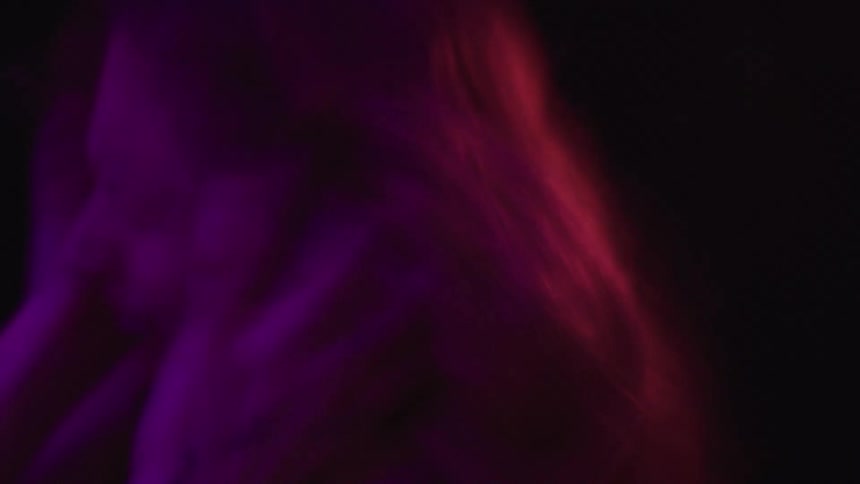 A blurry silhouette of a woman with long light colored hair is seen from the side as she brings her hands to her head and screams with a red light and a blue light flashing across her