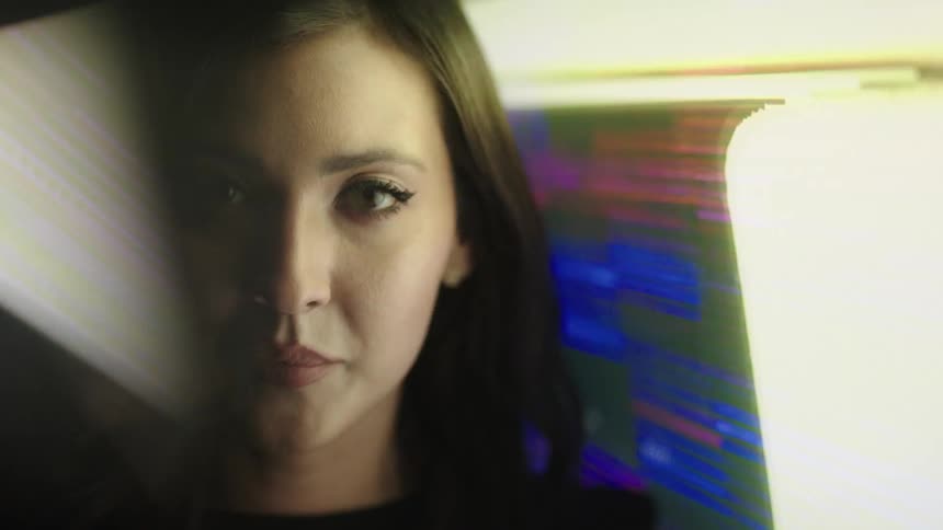 A womans face is seen in a close view with her long dark hair visible and her eyes looking directly into the camera as a colorful flashing screen illuminates her