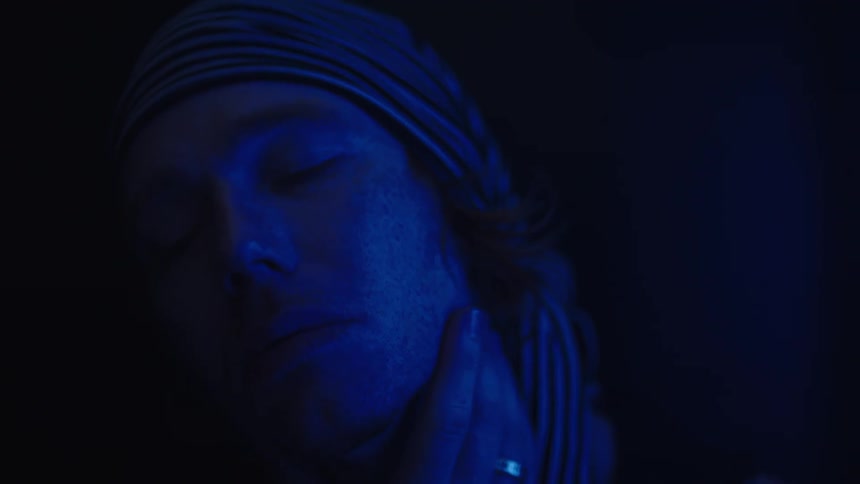 A persons face is shown in a very dark lowlit environment illuminated by a deep blue light they are wearing a striped bandana or head wrap and a hand with a ring on one finger is touching the side of their face