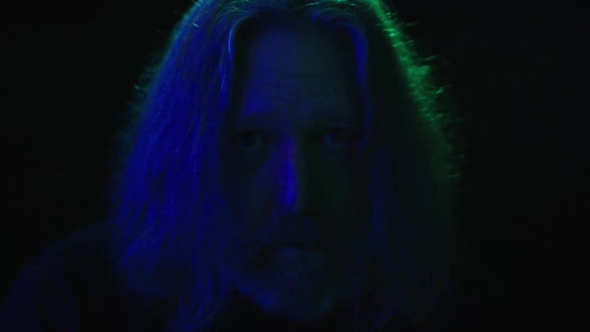 Longhaired man illuminated by blue and green light against a dark background