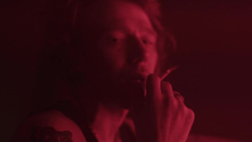 A man with light colored hair and a chain necklace smokes a lit cigarette with his face partially illuminated by a deep red light