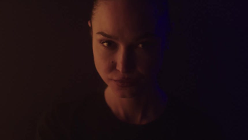 A young woman wearing a black tshirt sits in a dark room looking around curiously