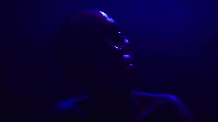 A dark silhouette of a woman with a shaved head and a bare chest is visible in a dark room