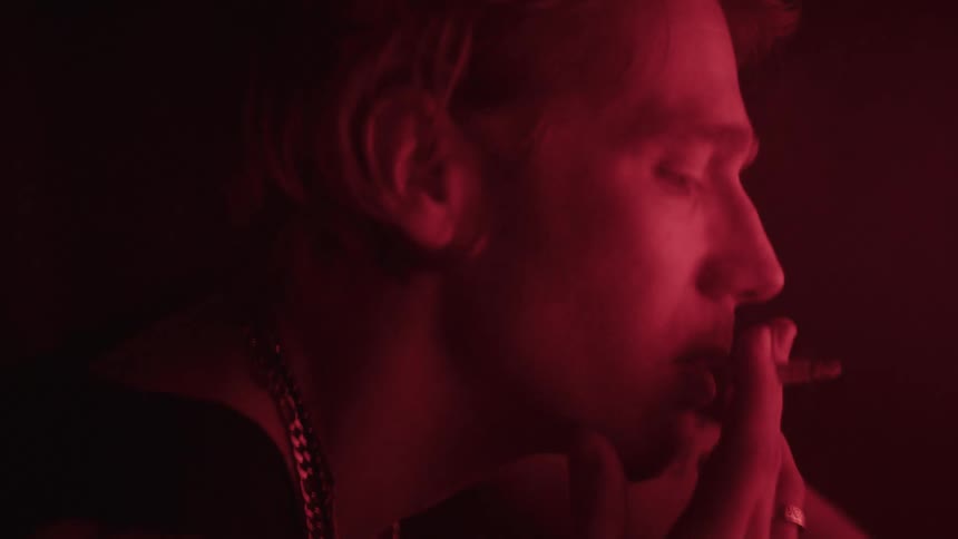 A closeup of a mans face in a lowlight redlit environment as he smokes a cigarette