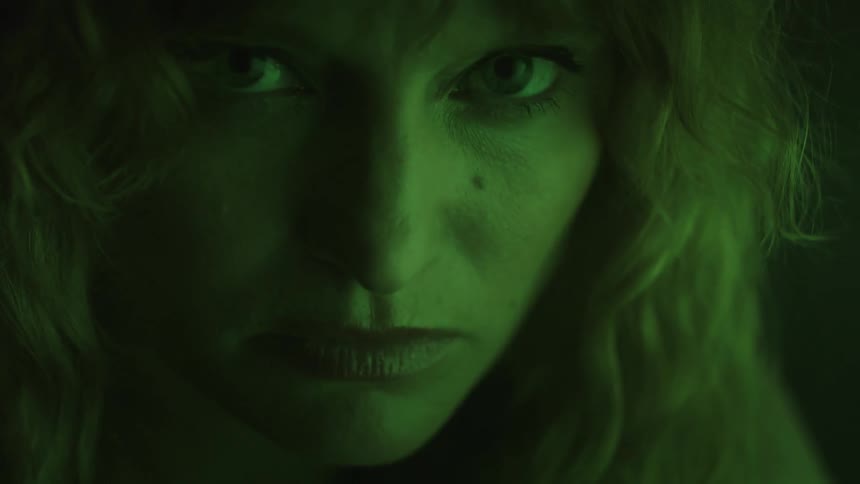 A womans face is shown in closeup under green light with a serious expression