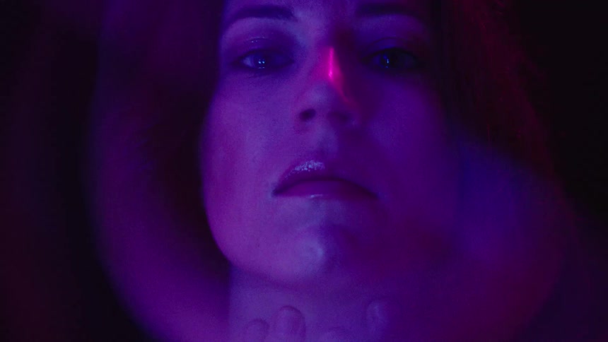 Womans face lit by purple and pink lights with a serious expression
