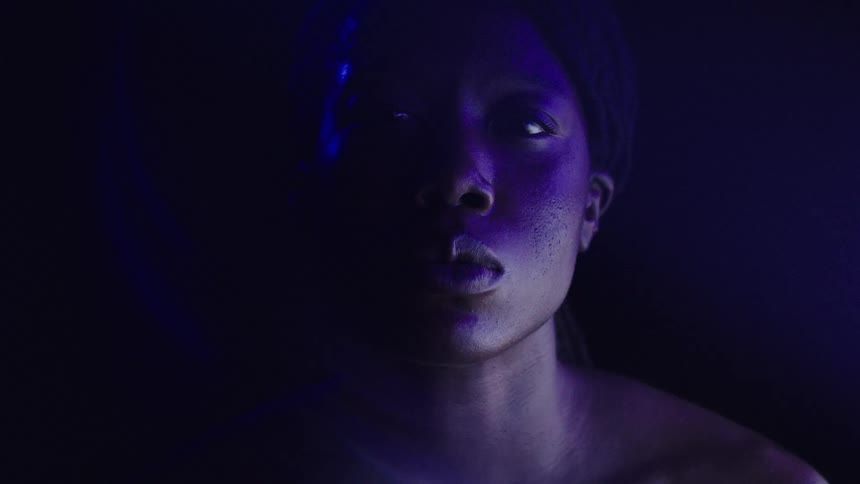 A young black woman shirtless sits in a dark room looking sad as a faint flickering light illuminates the atmosphere