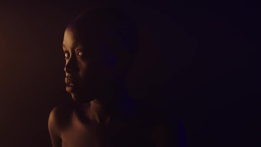 A woman with dark skin is seen in a close view with their head turning to look over her left shoulder while a warm orange light illuminates the left side of her face