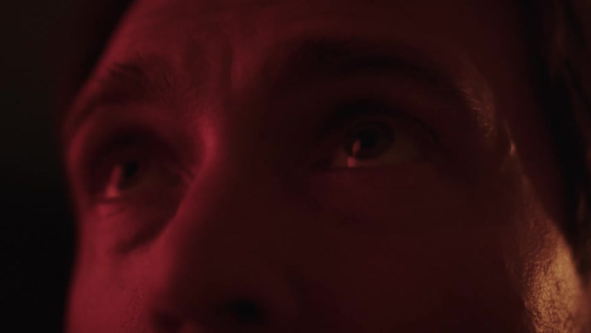 A man stands in a dark room illuminated by red light looking around with fear and apprehension