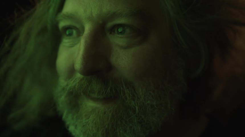 A bearded man with long hair looks ahead with a calm happy expression under greentinted lighting