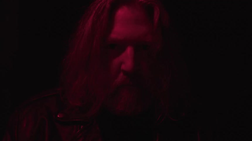 A man with long hair and a beard wearing a black jacket stands in a dark room illuminated by red light