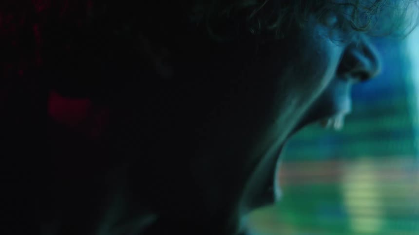 A young man stands facing a monitor screen screaming in anger at the distorted image displayed