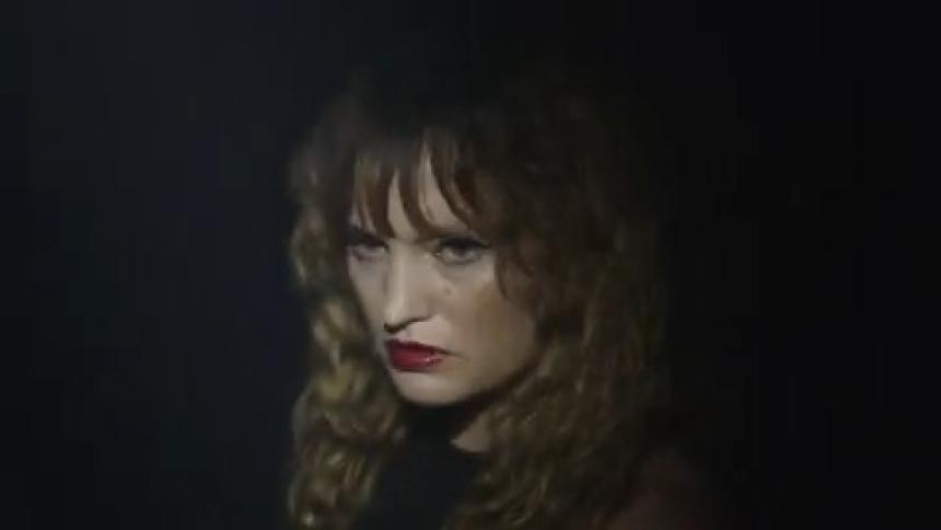 A young woman with blonde hair and a dark shirt stares directly at the camera with a neutral expression her lips painted with bright red lipstick under flashing lights