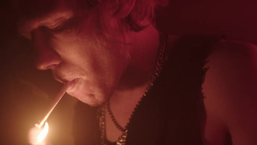 A close view shows a person with long light colored hair and a chain necklace smoking a cigarette as a lighter illuminates the cigarette and the persons face