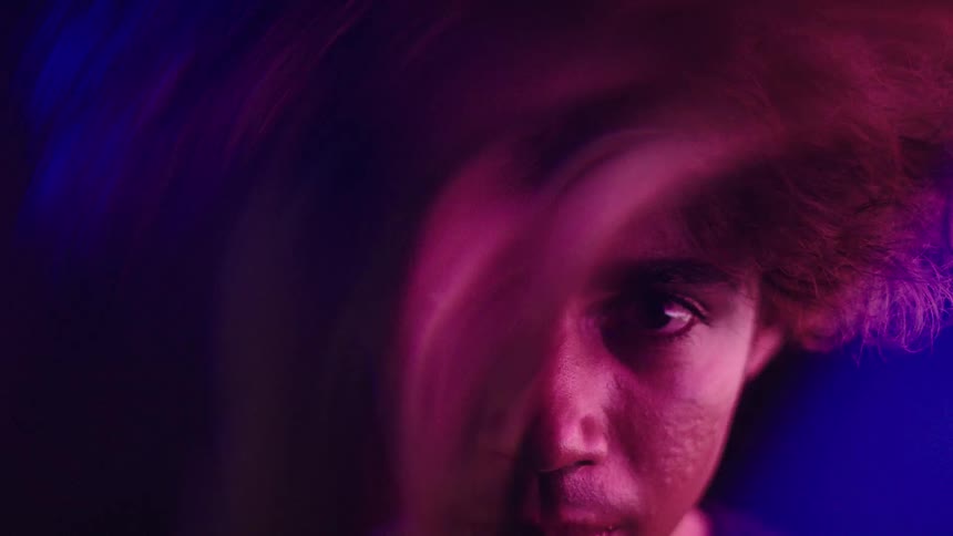 A mans face is seen in a close view illuminated by blue and purple light from the right side with his right eye open and his expression calm as he looks toward the camera
