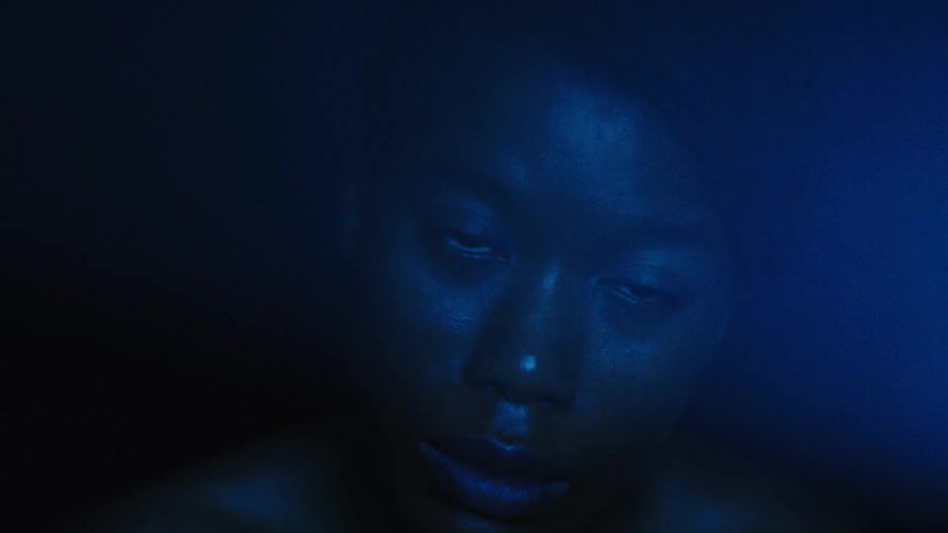 A close view shows a person with dark skin in a dark space with their face partially illuminated by a blue light as they move their head up and down and their eyes dart around