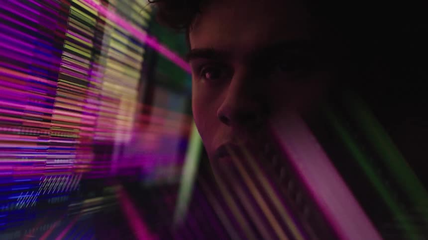 A persons face is illuminated by rapidly changing colored light as they look forward with their eyes opening and closing