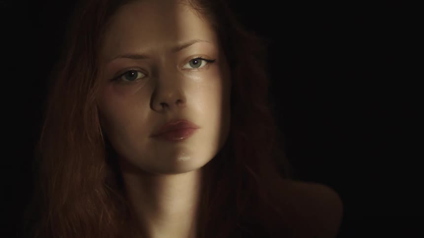 A young womans face is partially illuminated by a beam of light against a dark background