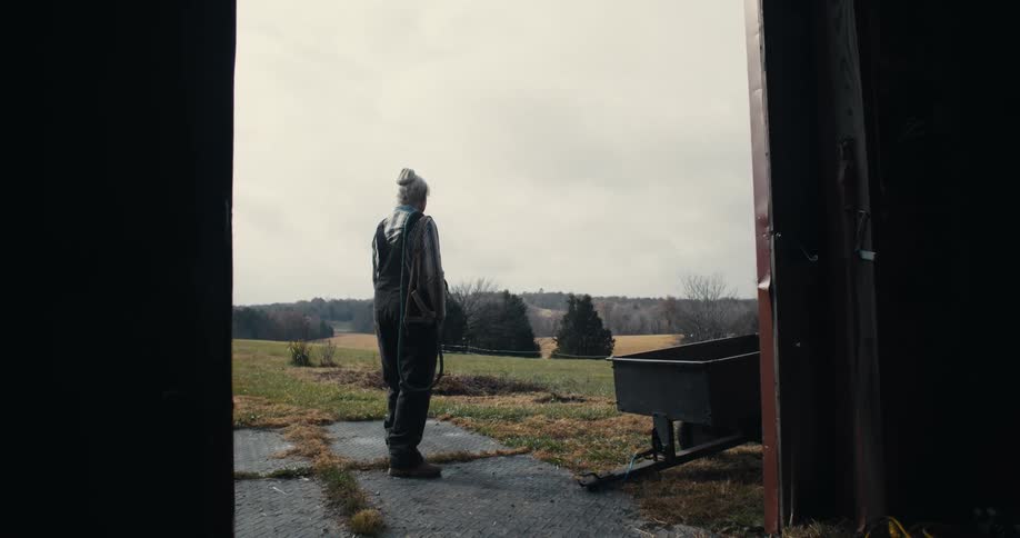 Elderly woman in work clothes looks at the field from the barn entrance