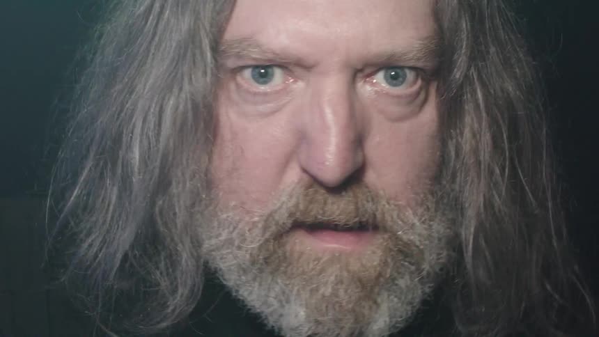 A man with long gray hair and a long gray beard looking directly into the camera with intense blue eyes under flashing lights